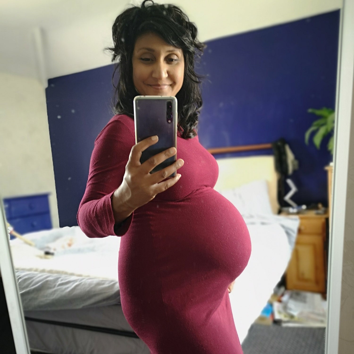 Simmi pregnant in a red dress taking a mirror selfie in a bedroom.