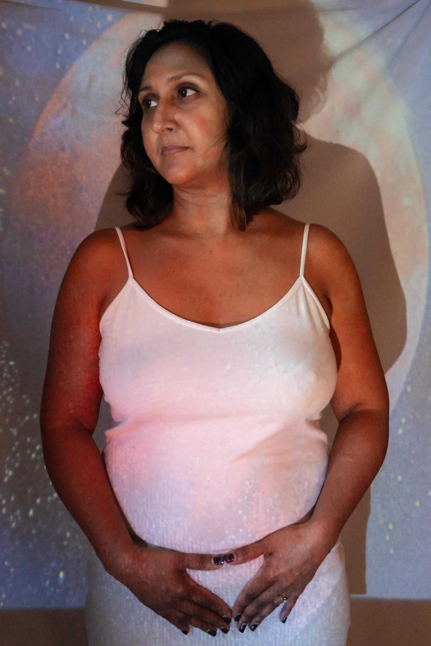Woman in contemplative pose with hands forming a triangle over her womb, with Mother Moon painting projected onto her.